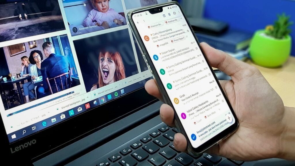 Hand holding a smartphone with an email inbox open beside a laptop, showing why email marketing for hotels must work on mobile